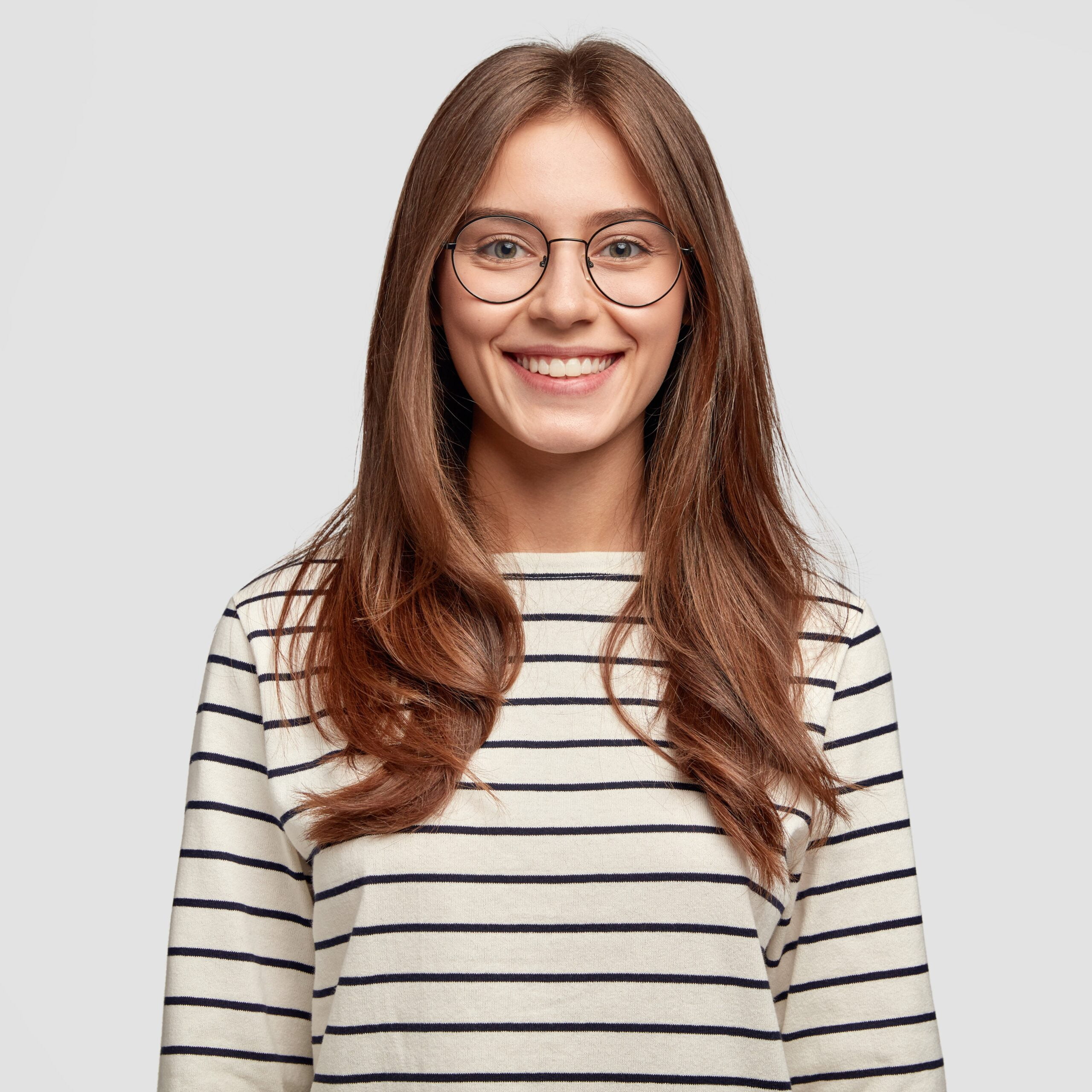Indoor shot of attractive European schoolgirl has toothy smile, dressed in striped sweater, rejoices positive moment in life, wears round spectacles. People, lifestyle and positive emotions concept